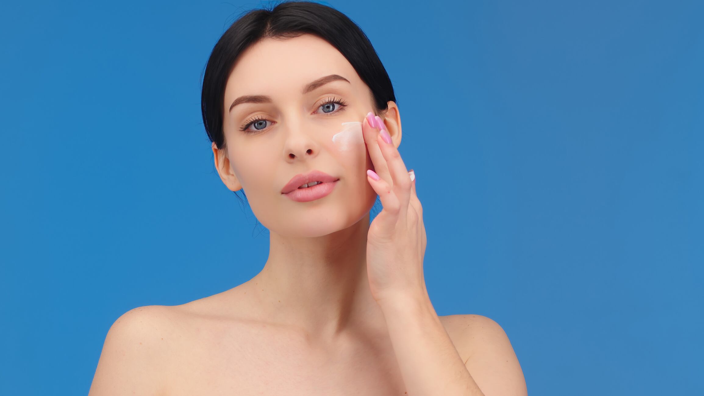 Face of a woman with blue eyes applying cream to her face and looking at the camera. Beautiful female model with perfectly clean fresh skin. Skin care or cosmetic advertising concept